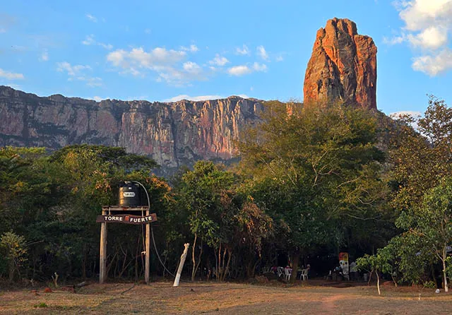 A tall, rocky monolith rises above a forest with a sign reading "Torre Fuerte" on a platform in the foreground in Bolivia.