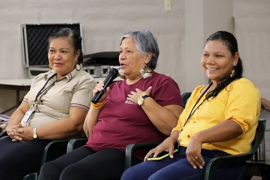 An Indigenous leader speaks into a microphone, flanked by two other Indigenous women, at a community gathering. The image highlights their engagement and representation in the discussion.