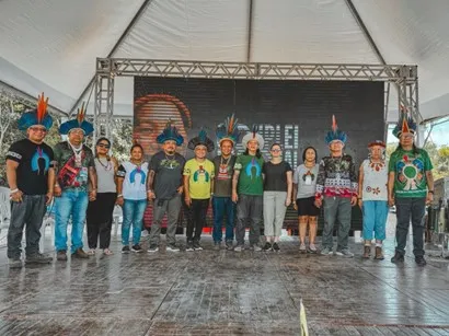 A group of Indigenous leaders, some wearing traditional headdresses, stand together for a photo on a stage in Brazil at an Indigenous network event.