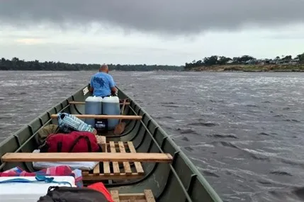 An Indigenous missionary travels by a long, narrow motorized boat down a wide, dark river under a cloudy, overcast sky.