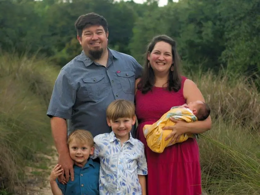 A smiling family of five. The father and two young boys stand beside the mother, who holds a baby.