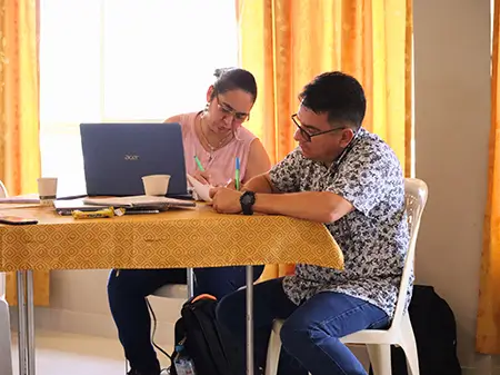 A woman and a man sit at a table working together with a laptop, writing notes on paper.