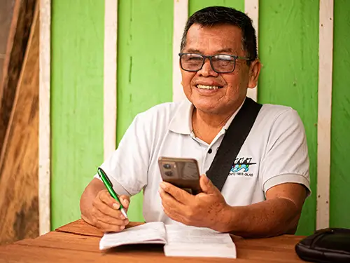 A smiling Indigenous man holding a smartphone and pen while writing in a notebook.