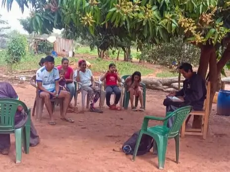 A leader conducting a community gathering with six people seated outdoors in a semicircle.