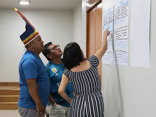 A woman points to a chart on a wall as two Indigenous leaders listen during a coaching session.