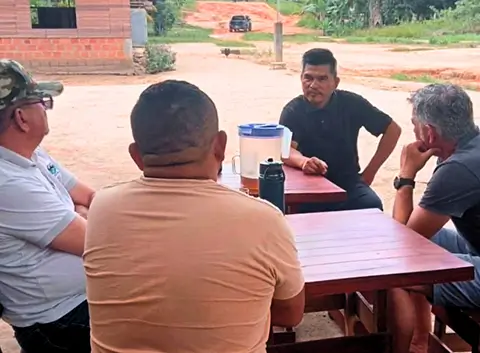 Four men sit around outdoor tables engaged in a conversation in a rural village setting.