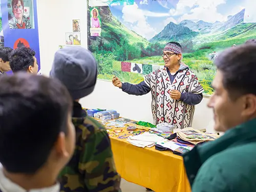 An Indigenous leader wearing traditional patterned clothing speaks to a small group gathered around a display table.