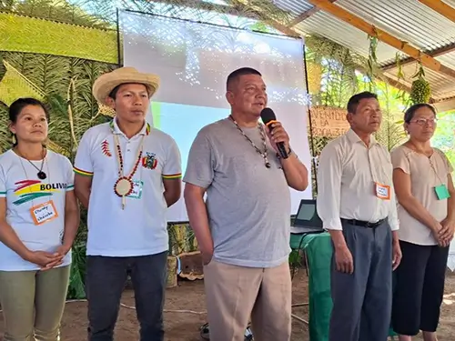 A group of five Indigenous leaders stands together as one man speaks into a microphone in front of a presentation screen.